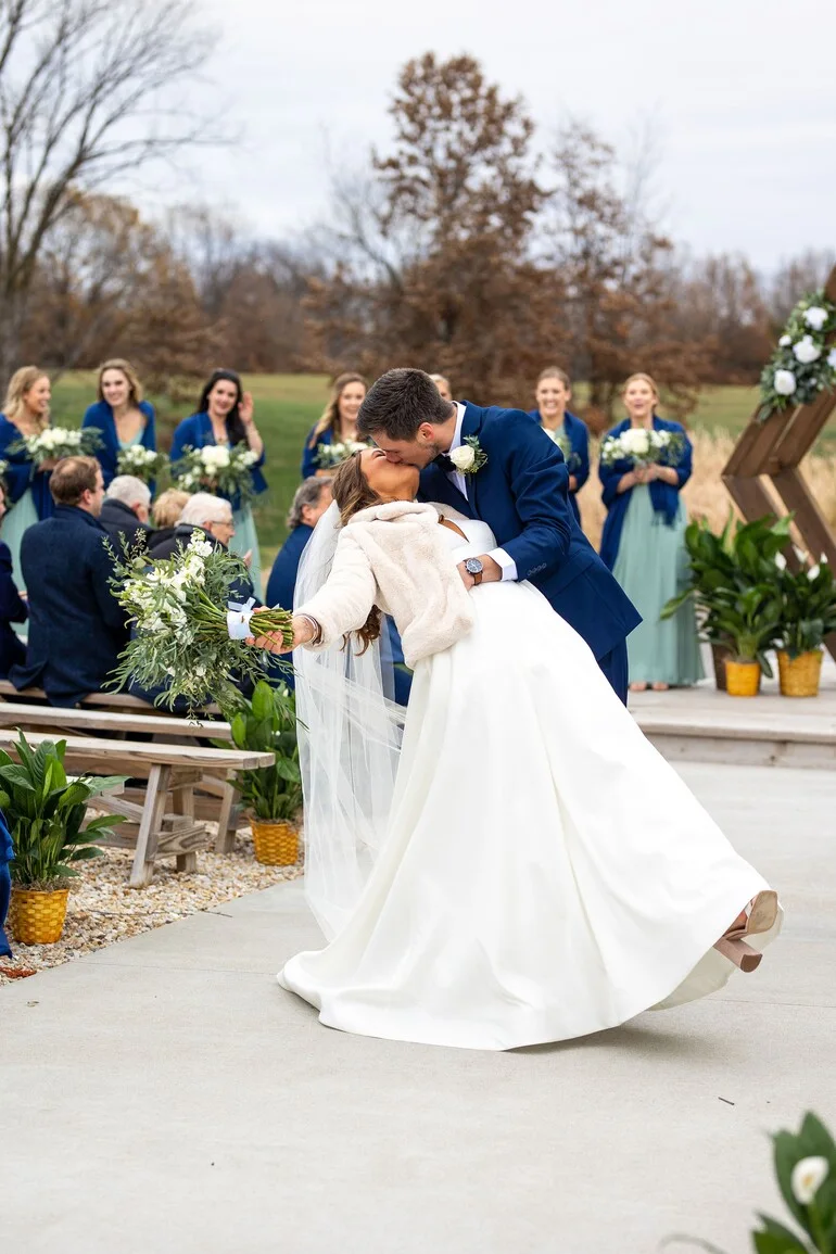 Bride and groom sharing a dip kiss at their outdoor wedding ceremony with the wedding party looking on