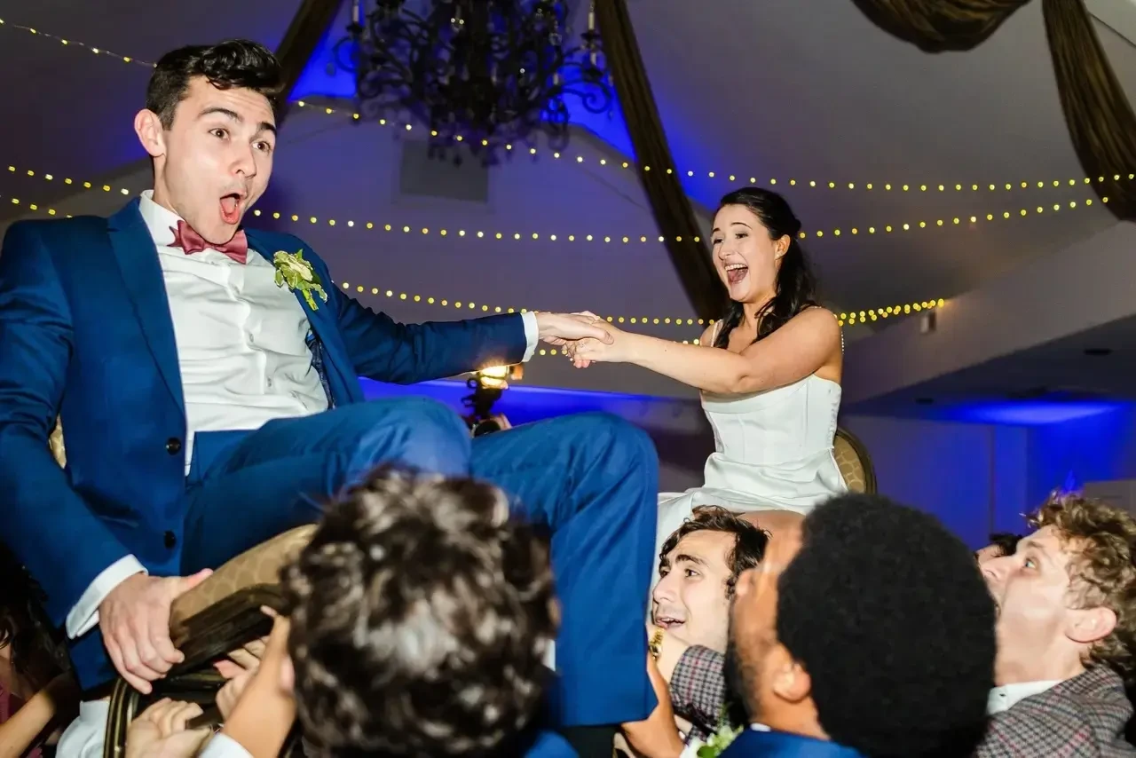 Bride and groom lifted in chairs reaching for each other during the hora at their wedding reception